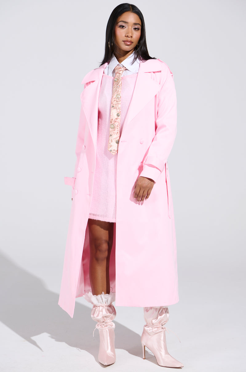 A woman models a chic ensemble with the PRETTY IN PINK TRENCH over a pink dress, effortlessly paired with a white shirt and floral tie. Her look is completed by pink high-heeled boots and white socks, set against a plain, well-lit backdrop.