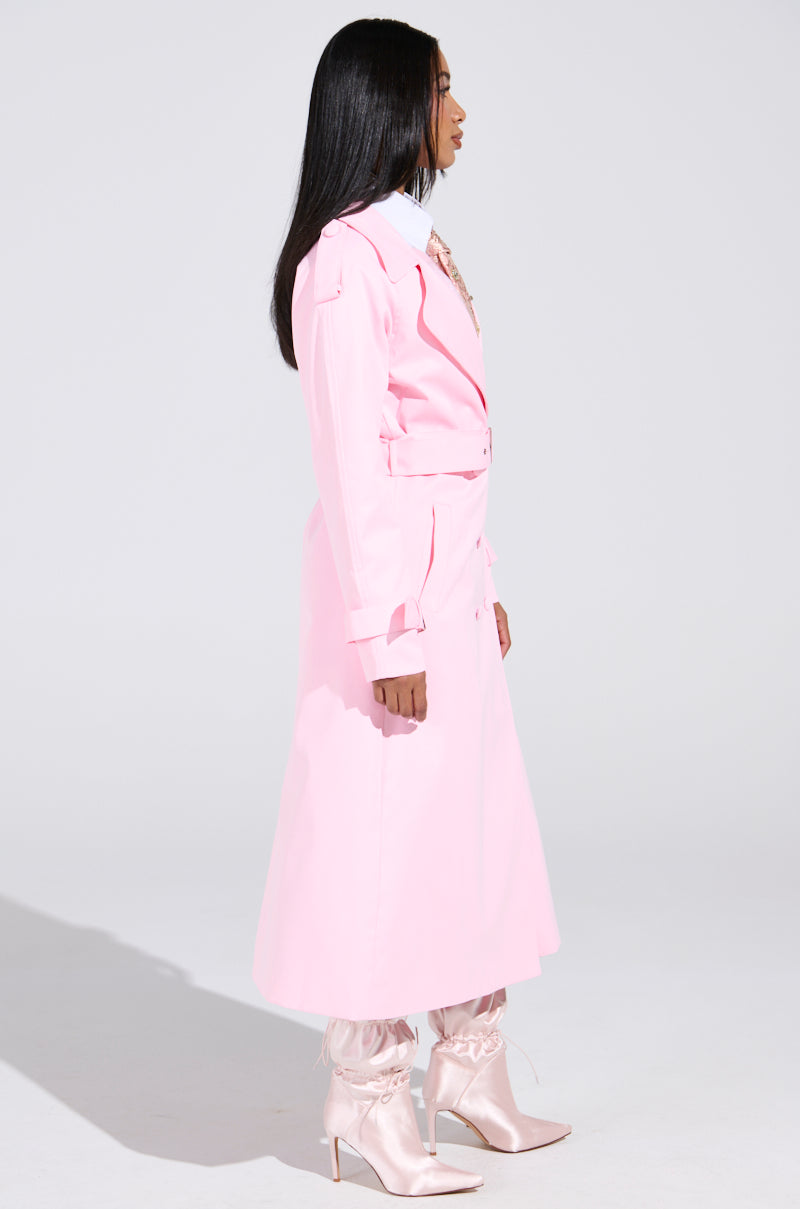 A person showcases the PRETTY IN PINK TRENCH in bubblegum pink, posed side profile with a belted waist and paired with matching satin boots, set against a plain white backdrop.