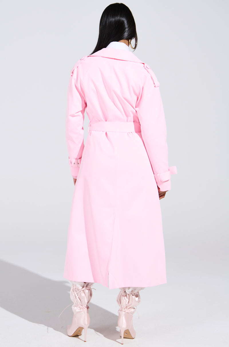 A person with long dark hair is facing away, wearing the PRETTY IN PINK TRENCH and matching high-heeled boots. The plain light-colored background highlights the vibrant elegance of this feminine spring ensemble.