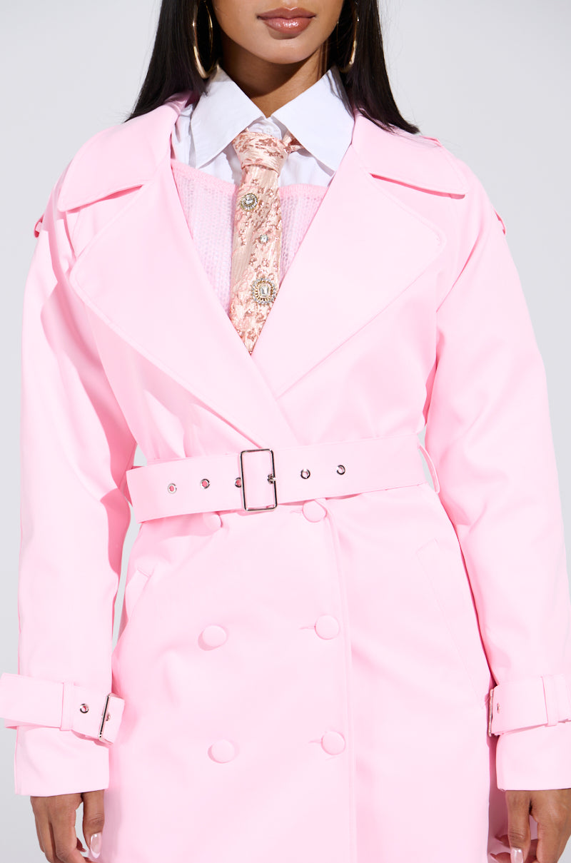 A person captivates in the PRETTY IN PINK TRENCH with a belt, buttons, and wide lapels, paired with a white collared shirt, floral pink tie, matching sweater, and hoop earrings that accentuate their long dark hair.