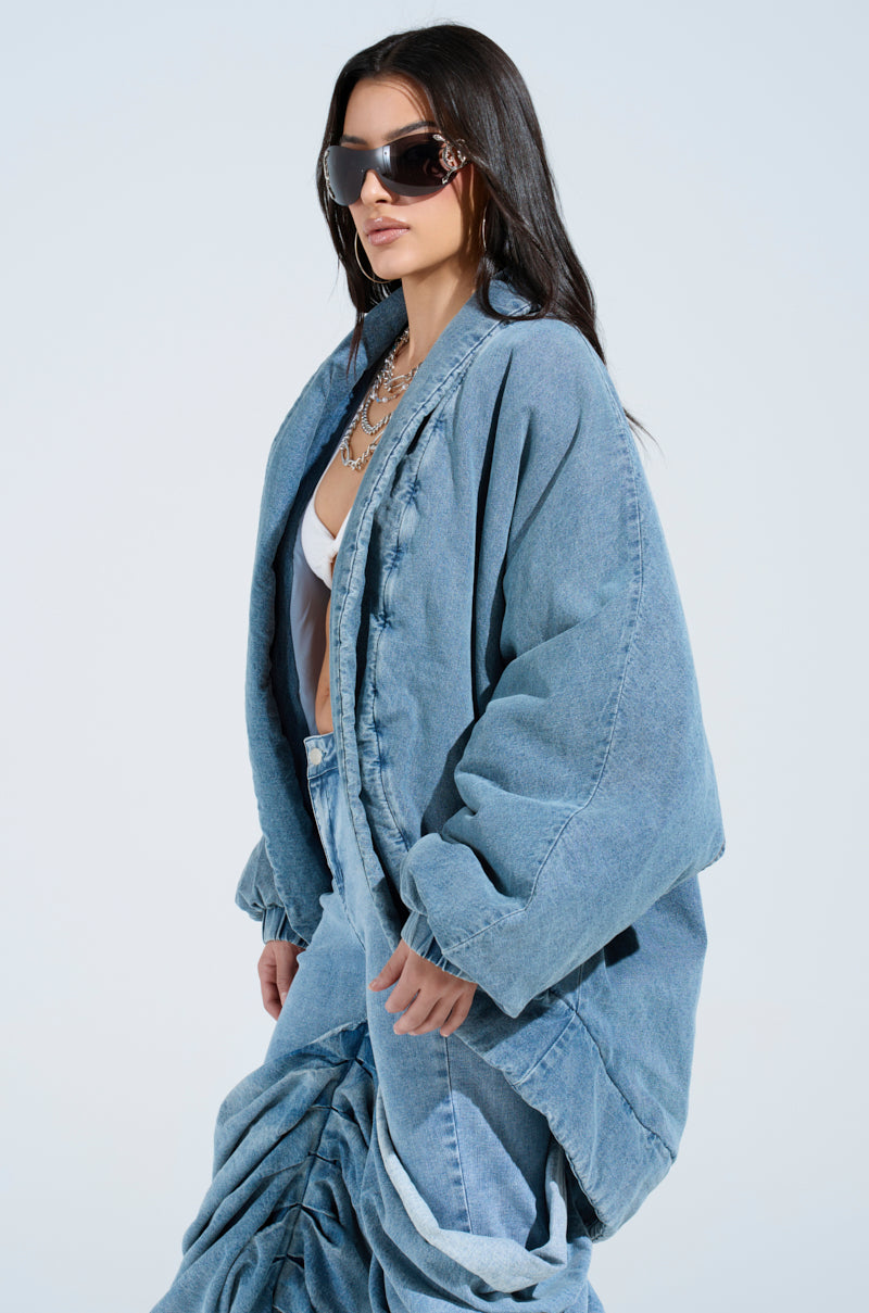 A person in a LAY UP AND CHILL DENIM COCOON COAT and long skirt stands against a plain background, sporting long dark hair, dark sunglasses, and layered necklaces for a stylish and contemporary look.