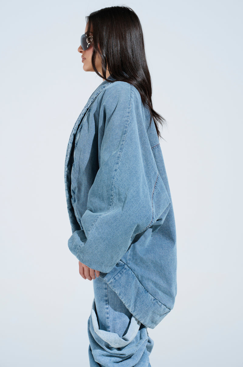A person with long dark hair wears a LAY UP AND CHILL DENIM COCOON COAT and pants, captured in profile, sporting sunglasses. The light blue background adds to the relaxed vibe of their look.