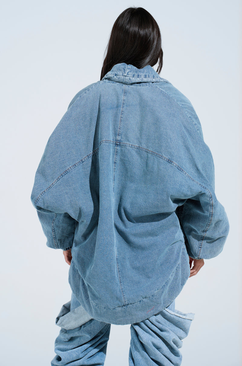 A person with long dark hair is wearing the LAY UP AND CHILL DENIM COCOON COAT, an oversized cotton denim piece, standing with their back to the camera against a light blue background.