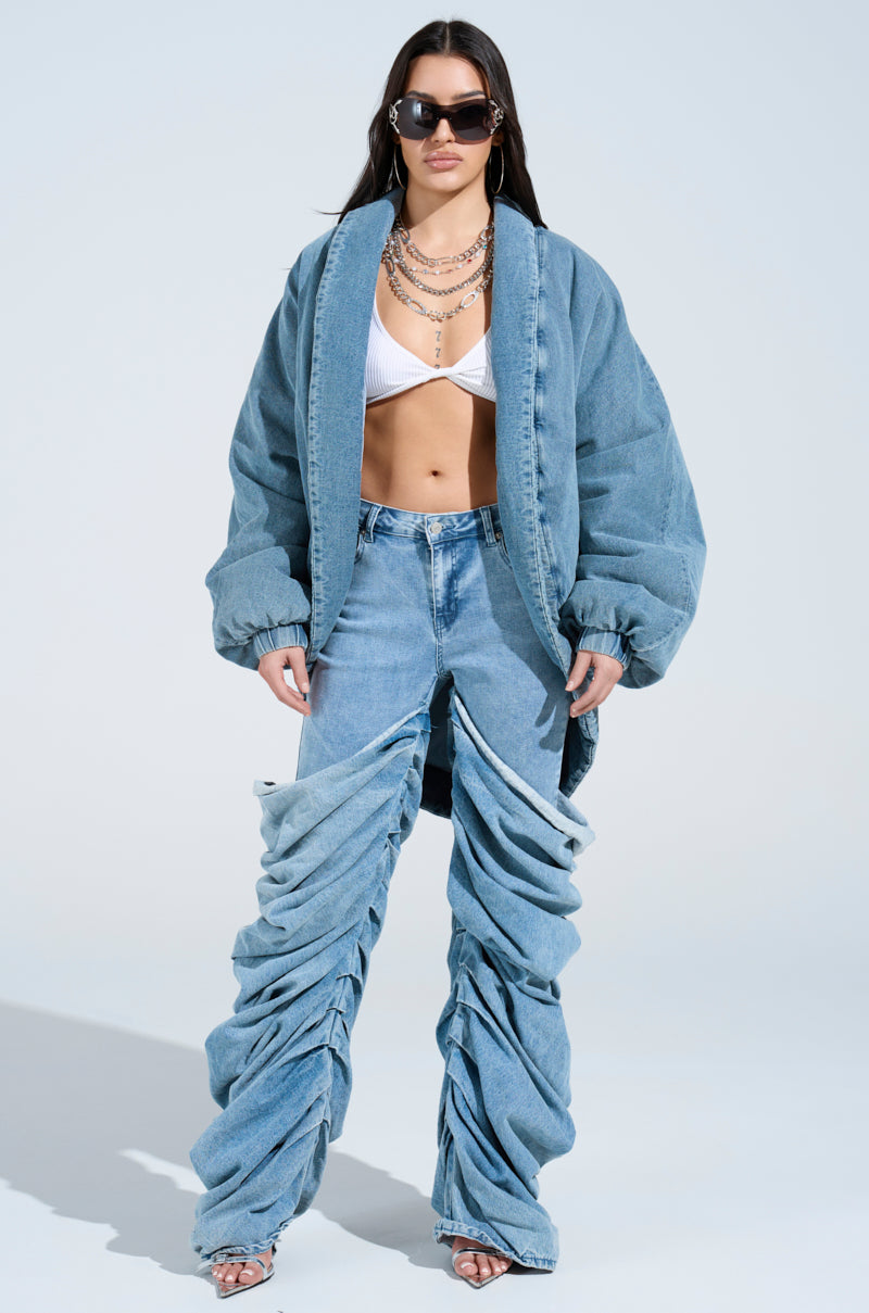 A person in a LAY UP AND CHILL DENIM COCOON COAT, white bralette, and distinctive ruched denim pants accessorizes with layered necklaces and sunglasses against a plain background.