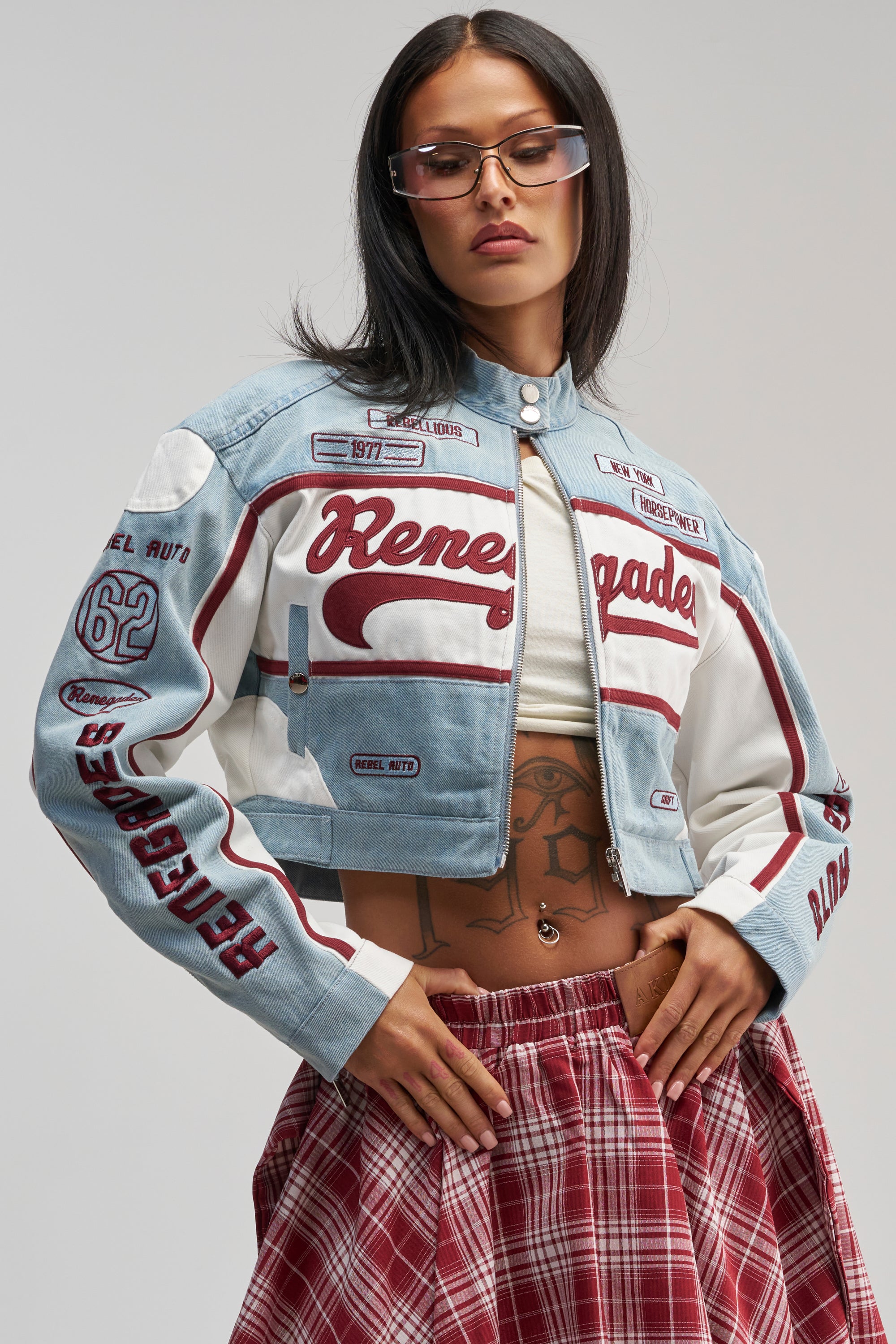 Wearing the VICTORY LAP DENIM MOTO jacket styled with a plaid red skirt, clear glasses, and a white top, a woman stands with one hand on her waist, revealing a torso tattoo against a plain background—a bold streetwear look featuring the jacket’s modern cropped silhouette.