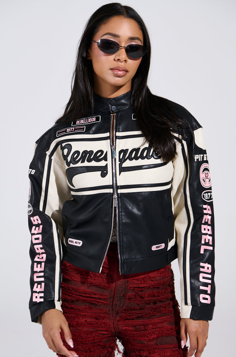 A person in the RENEGADES EMBROIDERED MOTO, a black and white faux leather jacket featuring auto racing patches, wears red textured pants and sunglasses. With long dark hair, they complete the bold color block look.