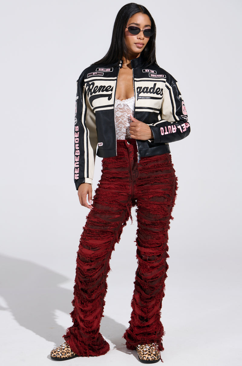 A woman poses against a plain backdrop wearing the RENEGADES EMBROIDERED MOTO jacket in black and white with text, red textured pants, leopard print shoes, and a lace top. Her sunglasses and flowing hair add to her confident look.