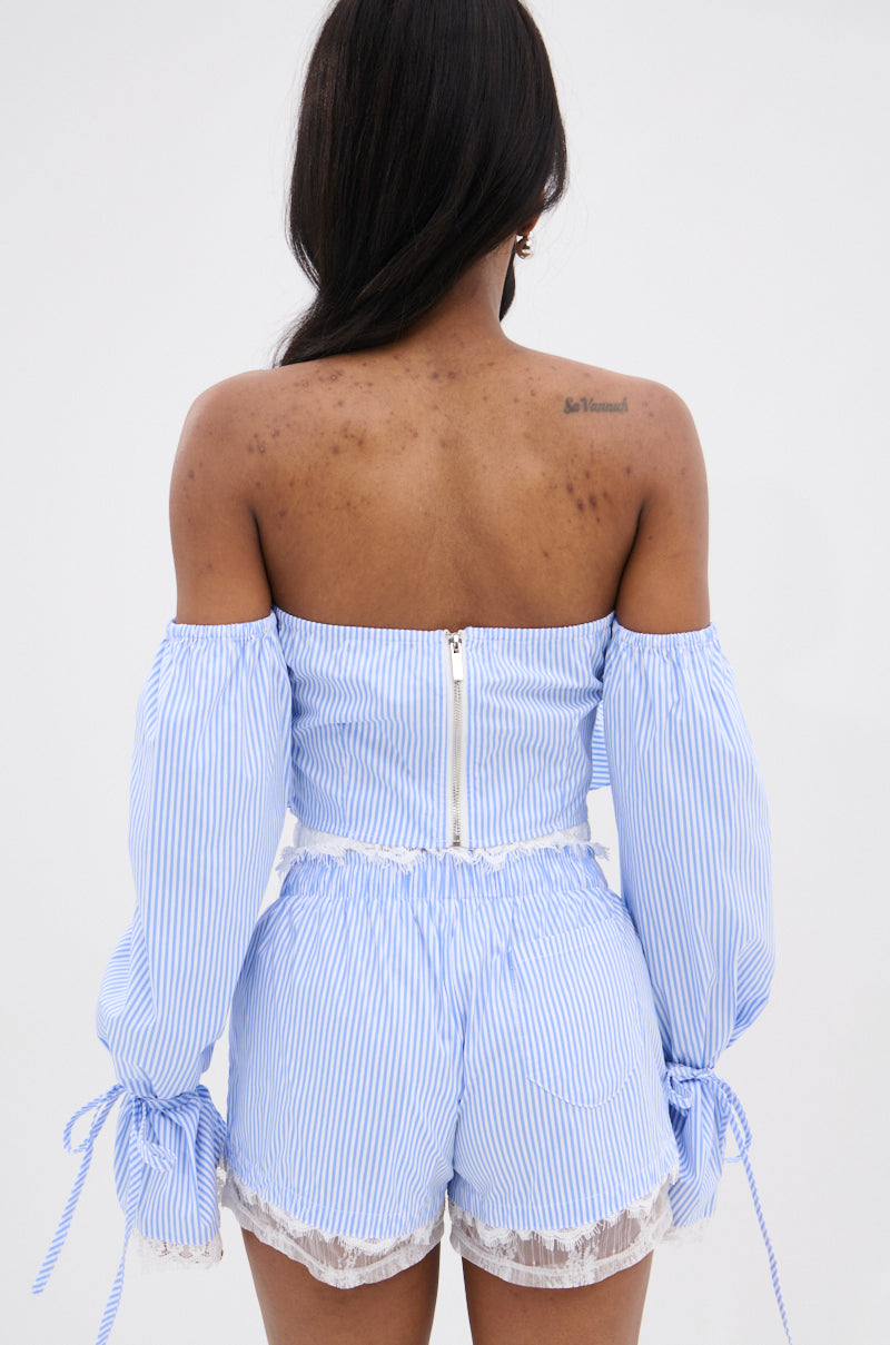 A woman with medium-toned skin and an upper back tattoo faces away, wearing the Prairie Chic Off the Shoulder Long Sleeve Blouse in light blue and white stripes.