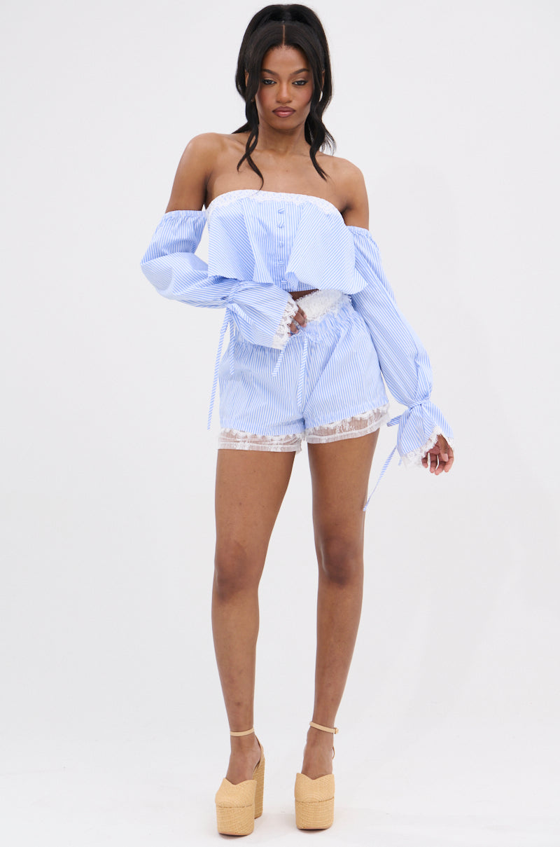 A woman stands against a white background wearing the PRAIRIE CHIC OFF THE SHOULDER LONG SLEEVE BLOUSE with blue and white stripes, paired with matching shorts featuring lace trim and beige platform heels.