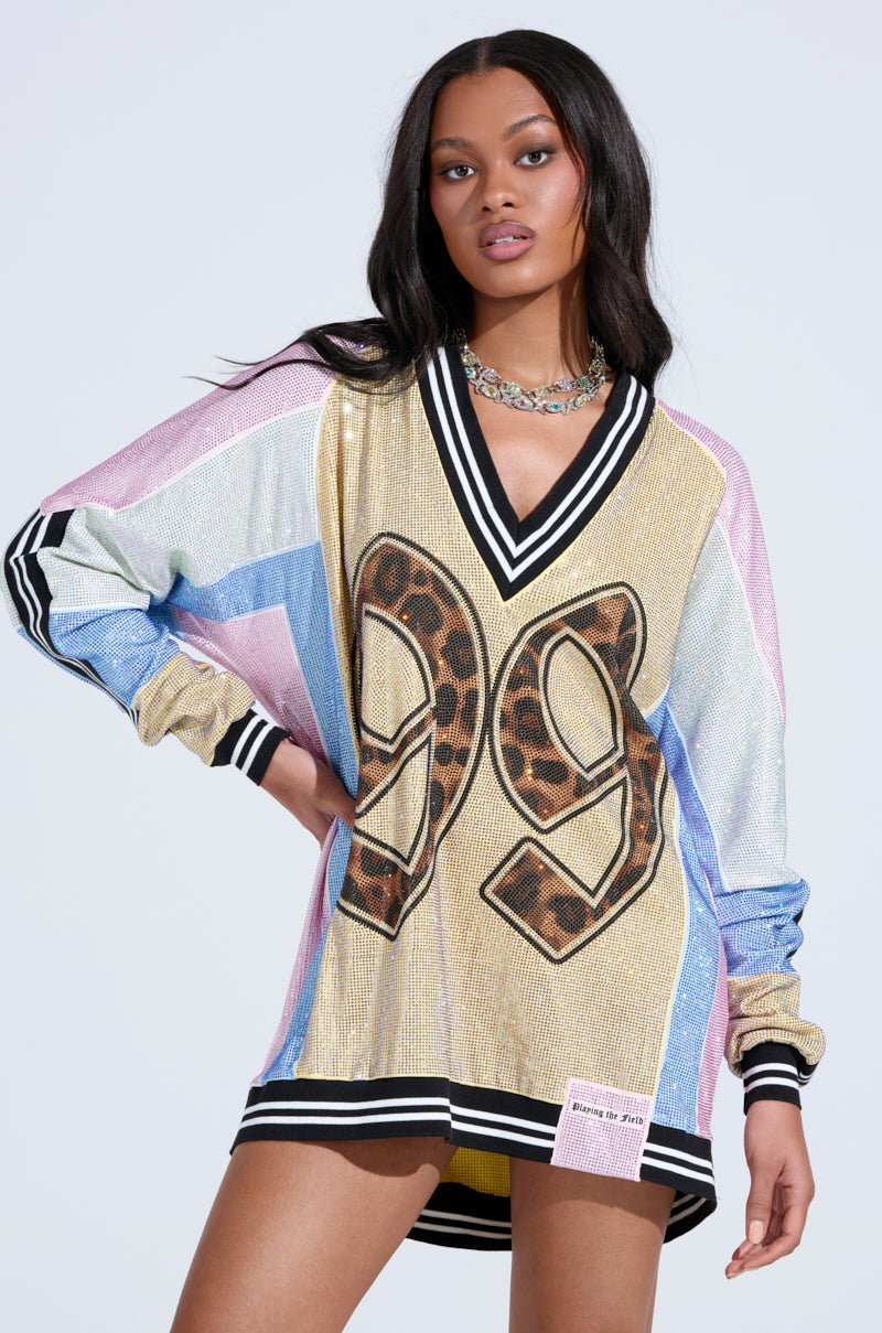 A woman wears the RYAN Colorblock Rhinestone Jersey Mini Dress in Pastel, a loose, long-sleeved dress with multicolored sequins, black and white trim, and "29" in leopard print. She has long wavy hair and a statement necklace.