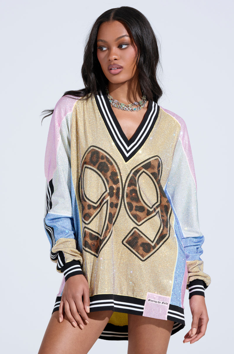 A woman in the RYAN Colorblock Rhinestone Jersey Mini Dress in pastel, featuring rhinestones, black and white striped cuffs and collar, and a bold leopard-print "99" on the front, looks to the side against a plain background.