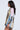 A woman with long wavy hair stands sideways in the RYAN Colorblock Rhinestone Jersey Mini Dress in Pastel, featuring a loose fit and black and white striped cuffs and hem. The background is plain and light-colored.