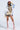 A woman in a pink cap, mesh veil, and the RYAN COLORBLOCK RHINESTONE JERSEY MINI DRESS IN PASTEL stands confidently. She pairs it with yellow gloves and tall lace-up pink platform boots against a plain white background.