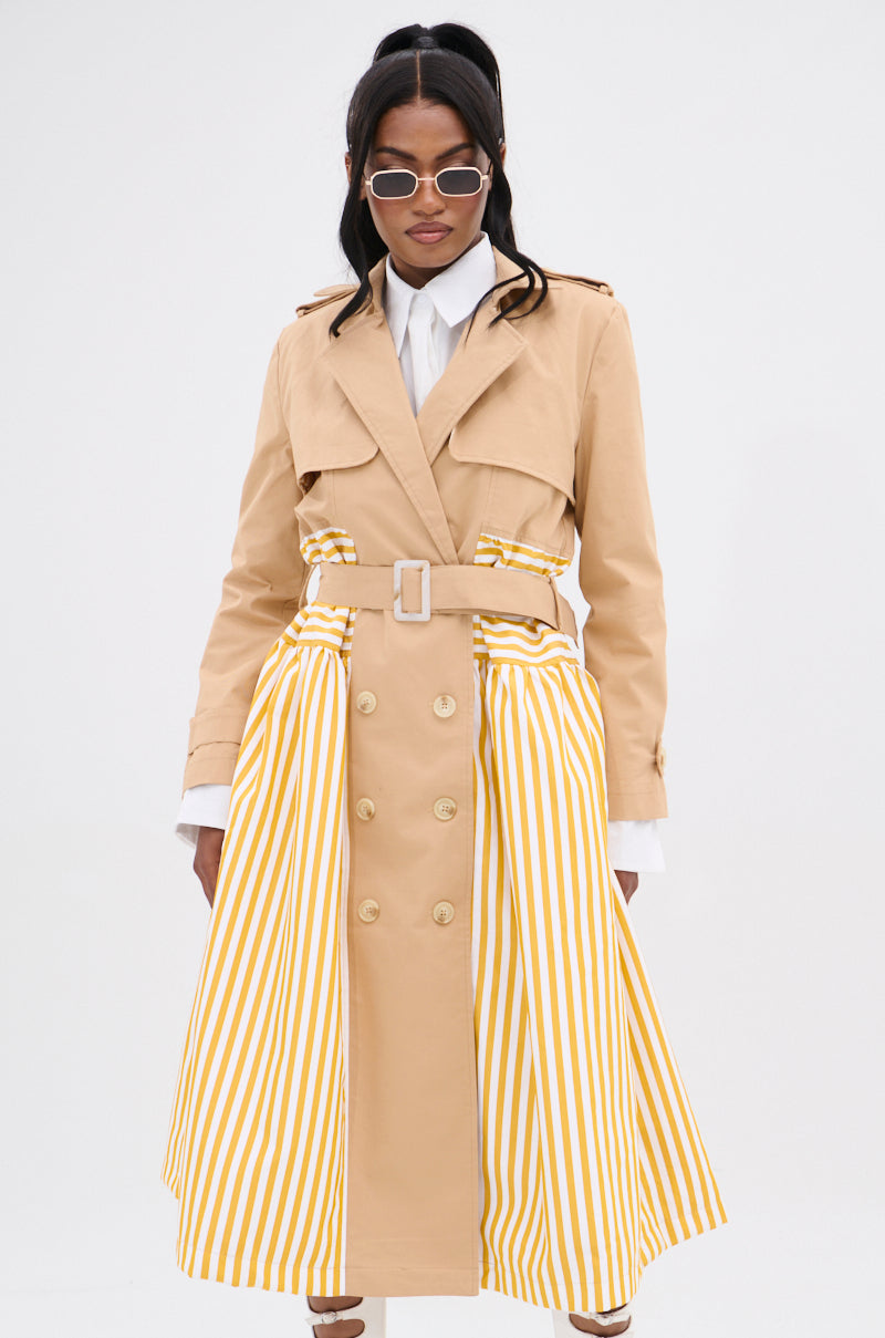 A person confidently models the LIKE A LADY STRIPED TRENCH, a double-breasted beige trench coat featuring yellow and white vertical striped panels. Paired with sunglasses and a white collared shirt, its unique design stands out against the plain white background.