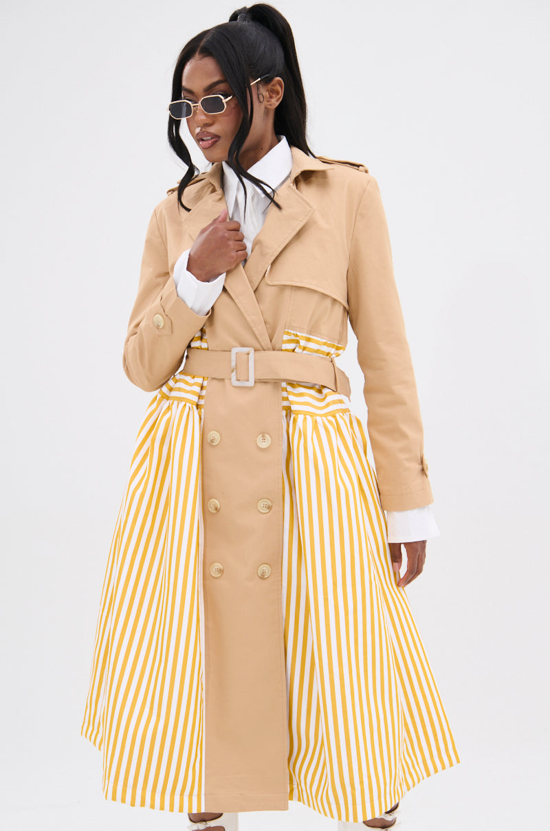 An individual with an hourglass figure models the LIKE A LADY STRIPED TRENCH, a beige coat with yellow and white striped panels, belted waist, and double-breasted design. They pair it with sunglasses and a crisp white shirt against a plain white backdrop.