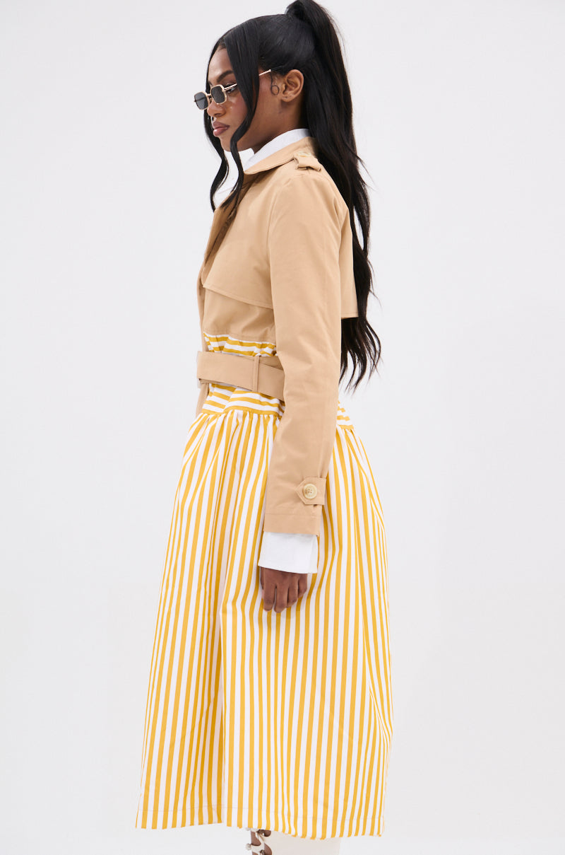 A woman in side profile embodies elegance in the LIKE A LADY STRIPED TRENCH, featuring a double-breasted silhouette over a yellow and white striped skirt. Her long hair is tied back with chic sunglasses, and the plain white background accentuates her graceful hourglass shape.