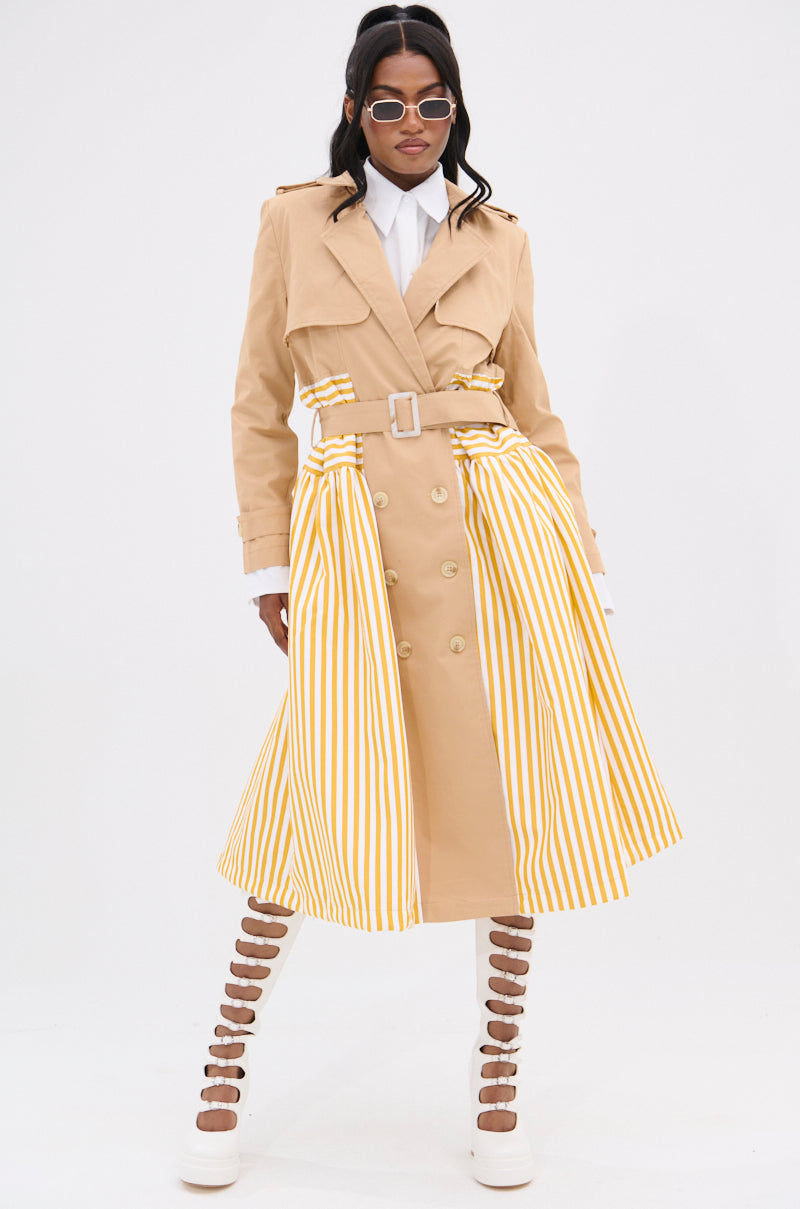 Wearing the LIKE A LADY STRIPED TRENCH, a person confidently poses in a tan double-breasted coat with yellow stripes, paired with a white shirt and strappy platform shoes. Sporting sunglasses and dark hair, they stand against a plain white background, highlighting an elegant hourglass shape.