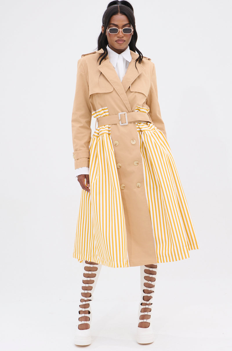 A woman stands confidently in the LIKE A LADY STRIPED TRENCH, showcasing its hourglass shape with yellow and white accents. She pairs it with a crisp white blouse, round sunglasses, and chic white platform shoes with cutout straps against a plain white backdrop.