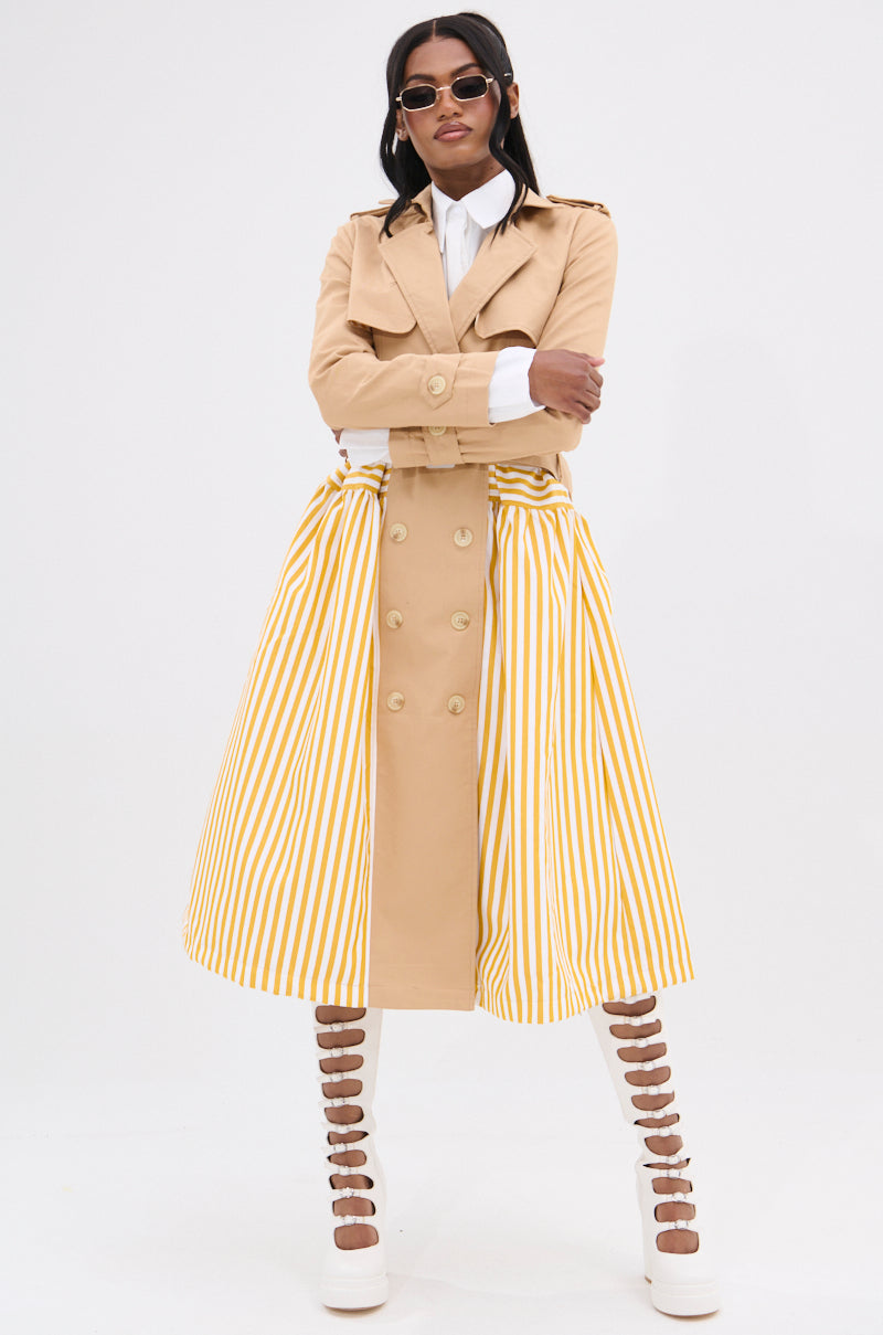 A person exudes confidence in the LIKE A LADY STRIPED TRENCH with a beige, double-breasted silhouette, paired with a yellow and white striped skirt. Long hair and sunglasses add flair, while tall, strappy white shoes enhance their shape against the plain white backdrop.
