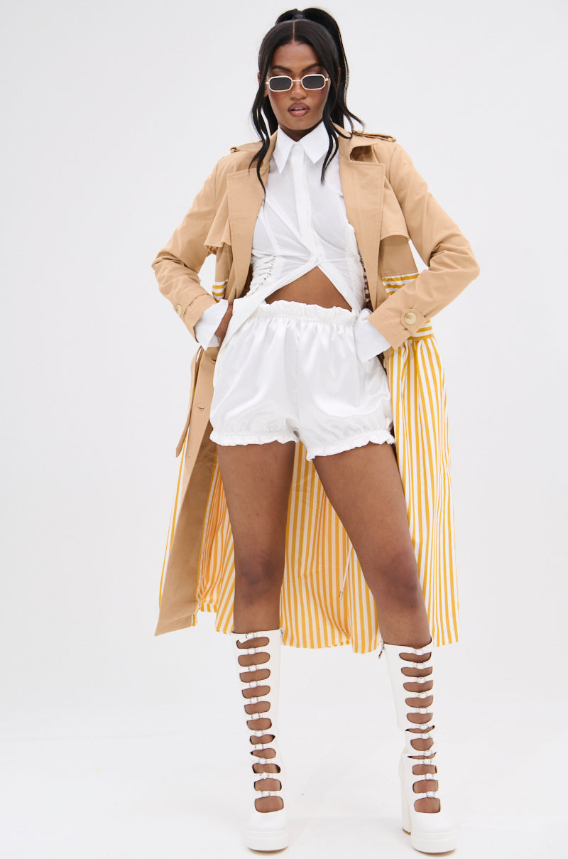 Confidently posing, the person dons the LIKE A LADY STRIPED TRENCH with a double-breasted look, paired with a white shirtdress and strappy white open-toed boots. They accessorize with sunglasses and place their hands on their hips for added flair.