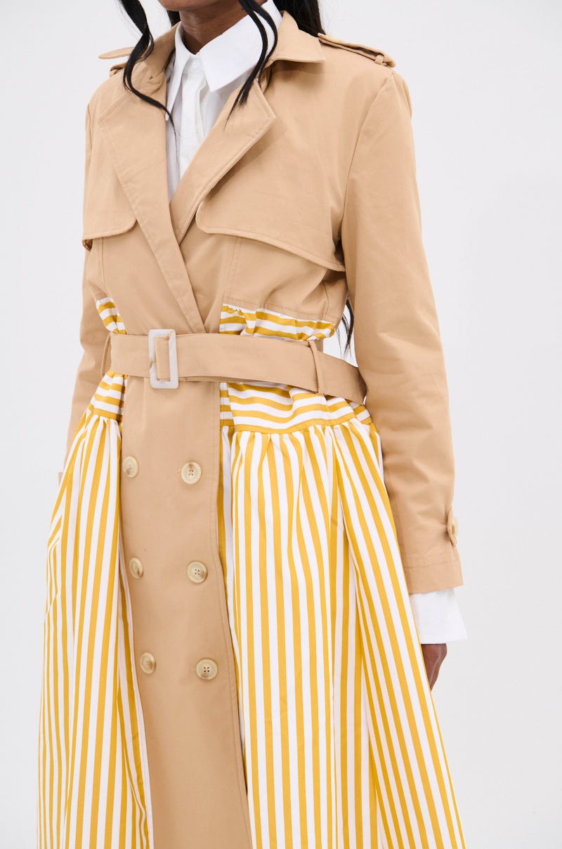 Wearing the "LIKE A LADY STRIPED TRENCH," a chic combination of beige and yellow vertical panels, a person accentuates an hourglass figure with belt and button details over a crisp white shirt against a plain white backdrop.