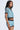 A young woman with long straight hair models the REBEL COLLARED TOP featuring a bold blue, brown, and white zigzag pattern with embroidered patches, paired with matching shorts. She poses against a plain light backdrop.