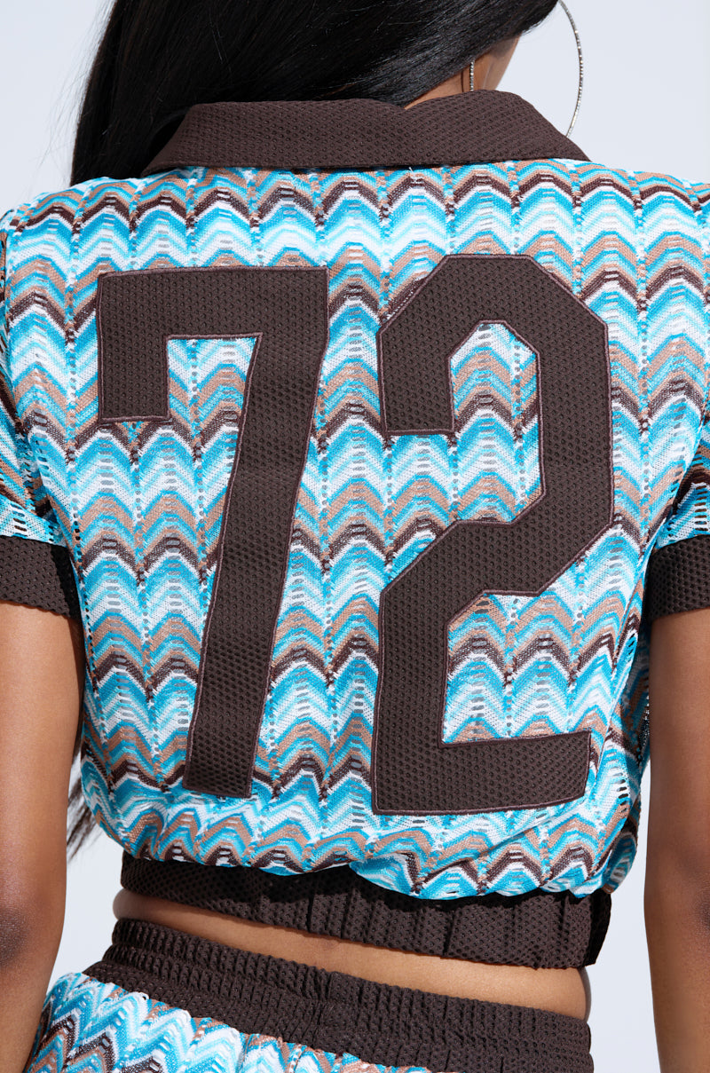 Someone wears the REBEL COLLARED TOP, a textured blue, brown, and white shirt with a colorful zigzag pattern, short sleeves, and a large dark brown 72 on the back. Only the upper back and part of the waist are visible.