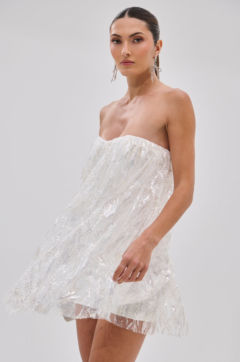 A woman with slicked-back hair poses confidently against a light gray background, wearing the SHINE BRIGHT SEQUIN MINI DRESS—strapless, white, covered in sparkling sequins and silver beads—finished with dangling earrings.