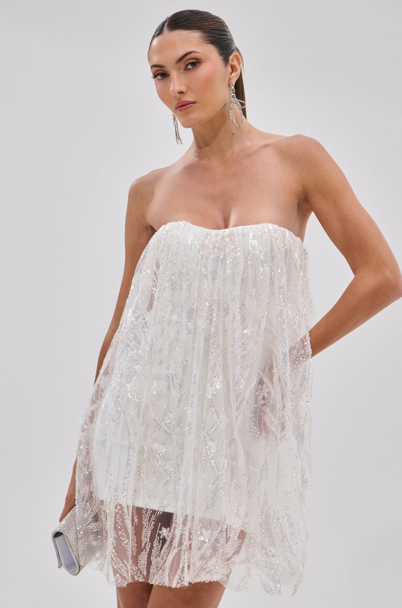 A woman in the SHINE BRIGHT SEQUIN MINI DRESS with a strapless, white sheer overlay stands against a plain light background, holding a silver clutch and wearing long earrings with slicked-back hair.