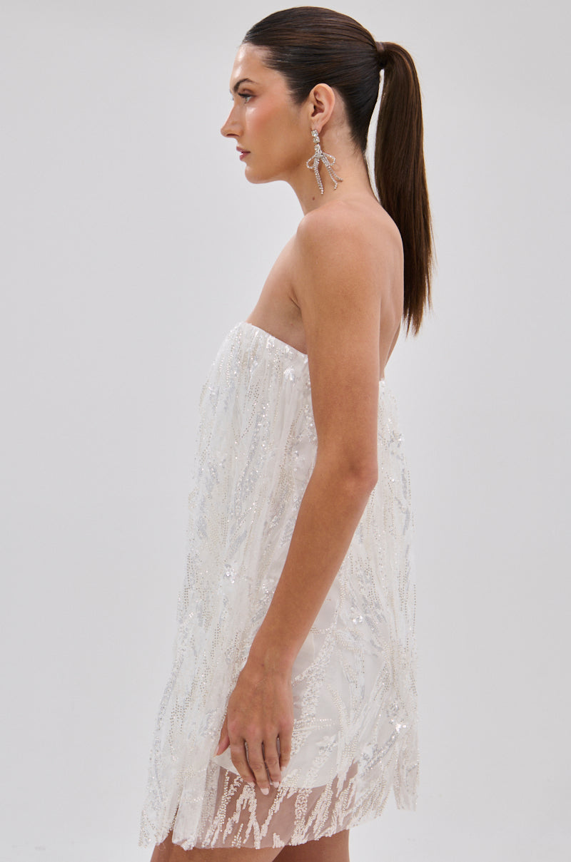 A woman with long, straight brown hair in a ponytail wears the SHINE BRIGHT SEQUIN MINI DRESS and statement earrings, standing in profile against a plain light gray background.