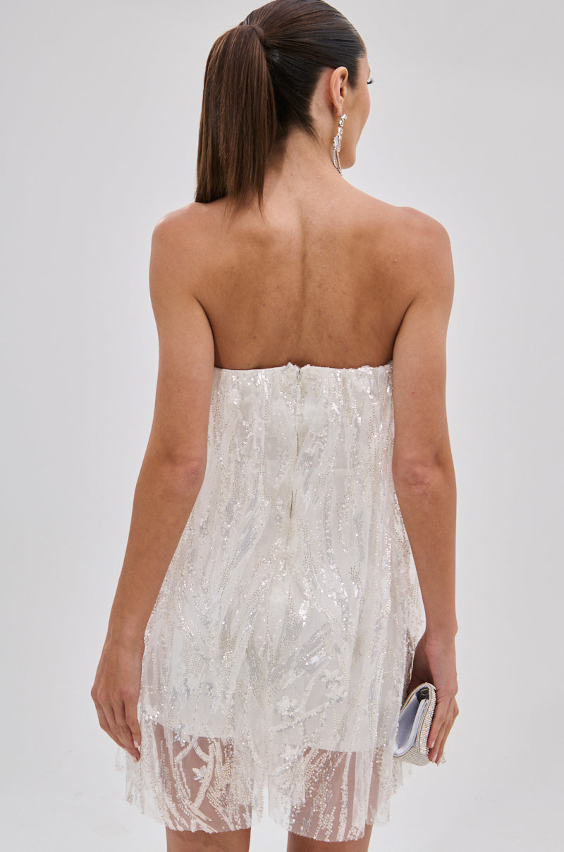 A woman with straight brown hair in a ponytail wears the SHINE BRIGHT SEQUIN MINI DRESS, a strapless white sequin mini dress. Holding a small silver clutch, she faces away to reveal the sparkling back of her dazzling outfit.