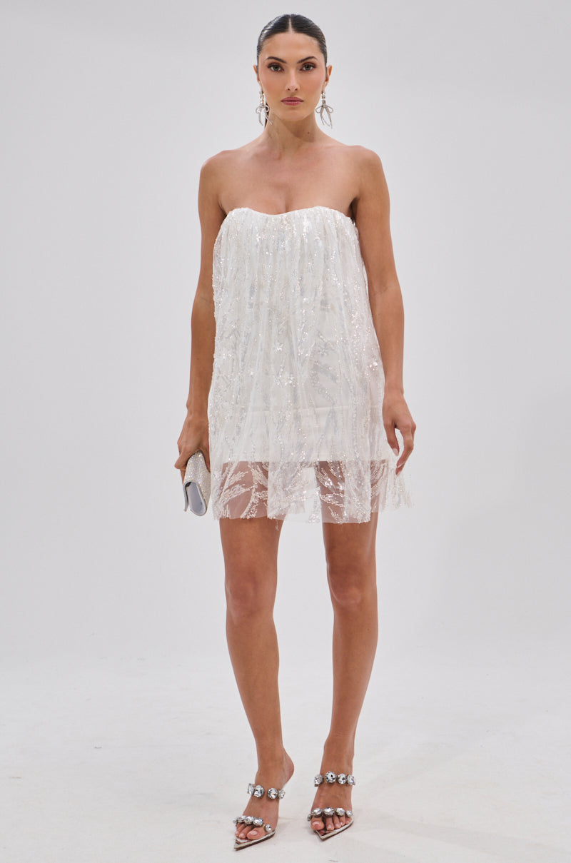A woman wears the SHINE BRIGHT SEQUIN MINI DRESS with a sheer hem, silver high-heeled sandals, drop earrings, and a small silver clutch while standing against a plain white background.