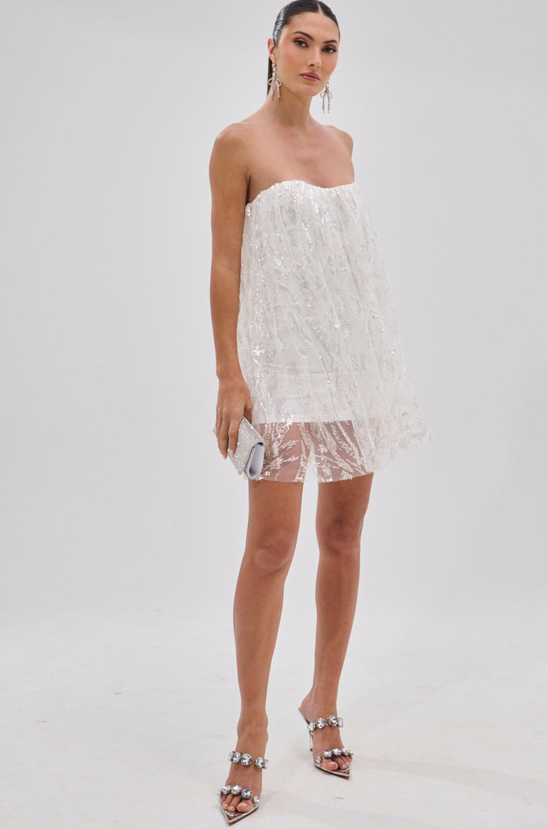 A woman stands against a plain white background in the SHINE BRIGHT SEQUIN MINI DRESS with a sheer lace hem, silver heeled sandals with jeweled accents, drop earrings, and holding a small silver clutch.