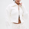 A person with short dark hair and visible tattoos stands against a white background, wearing the SEE ME COMING JACQUARD MOTO jacket with high-waisted, silver-studded shorts and large geometric earrings.
