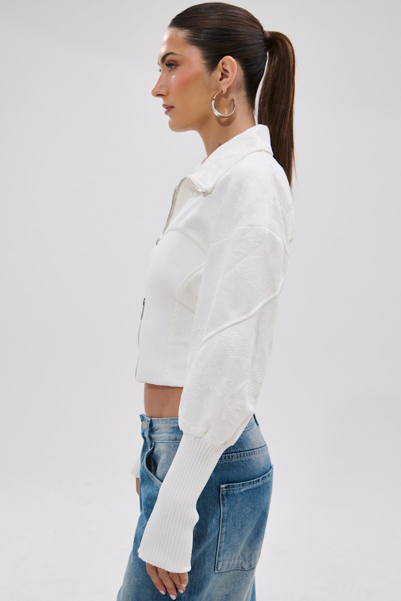 A woman with long brown hair in a ponytail, gold hoop earrings, and blue jeans wears the SEE ME COMING JACQUARD MOTO jacket against a plain light background.