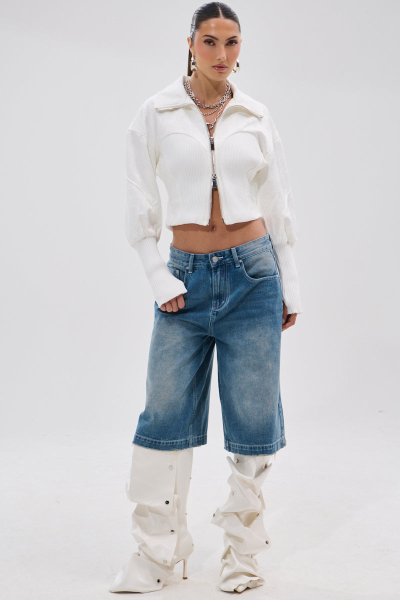 A woman with slicked-back hair poses against a plain background in the SEE ME COMING JACQUARD MOTO jacket, knee-length wide denim shorts, layered necklaces, and tall white slouchy heeled boots.
