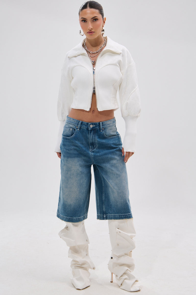 A woman stands against a plain background in the SEE ME COMING JACQUARD MOTO white cropped jacket, long denim shorts, layered necklaces, and slouchy white heeled boots. Her slicked-back hair and confident pose complete the look.