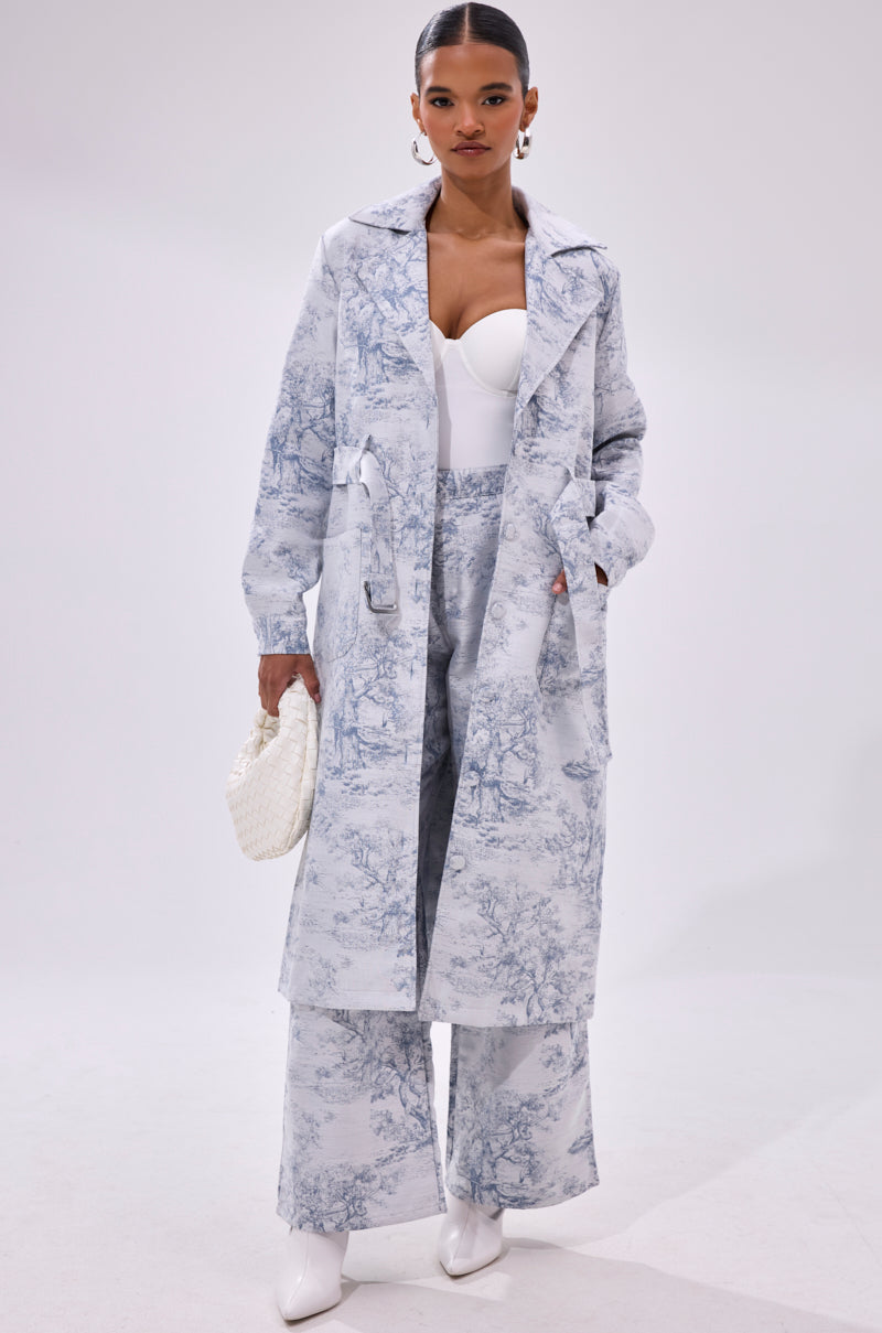 A woman models the BOOKED N BUSY JACQUARD TRENCH, a light blue and white scenic toile print long coat, paired with matching wide-leg pants, a white bustier top, heeled boots, and a woven handbag against a plain background.