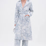 A woman stands against a white background wearing the BOOKED N BUSY JACQUARD TRENCH, styled with matching wide-leg pants, white heeled sandals, silver hoop earrings, and a small white handbag.