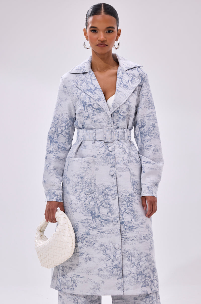 A woman wears the BOOKED N BUSY JACQUARD TRENCH with matching pants and large hoop earrings, holding a white woven handbag against a plain white background.
