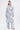 A woman wears the AKIRA Label BOOKED N BUSY JACQUARD TRENCH with matching wide-leg pants, white heeled sandals, hoop earrings, and a small white handbag while standing against a plain white background.