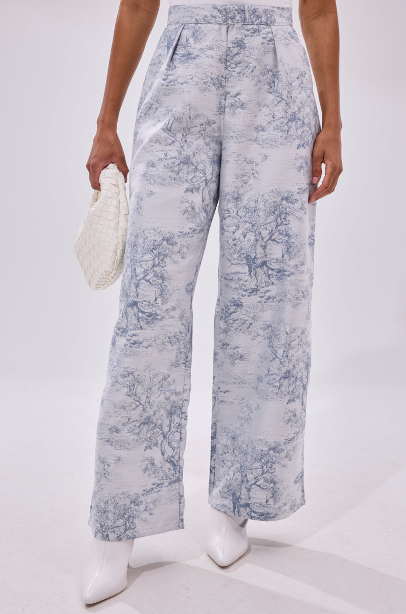 Someone wears the BOOKED N BUSY JACQUARD TROUSER, featuring a blue and white scenic print, styled with white heeled boots and a woven white clutch against a plain white background.