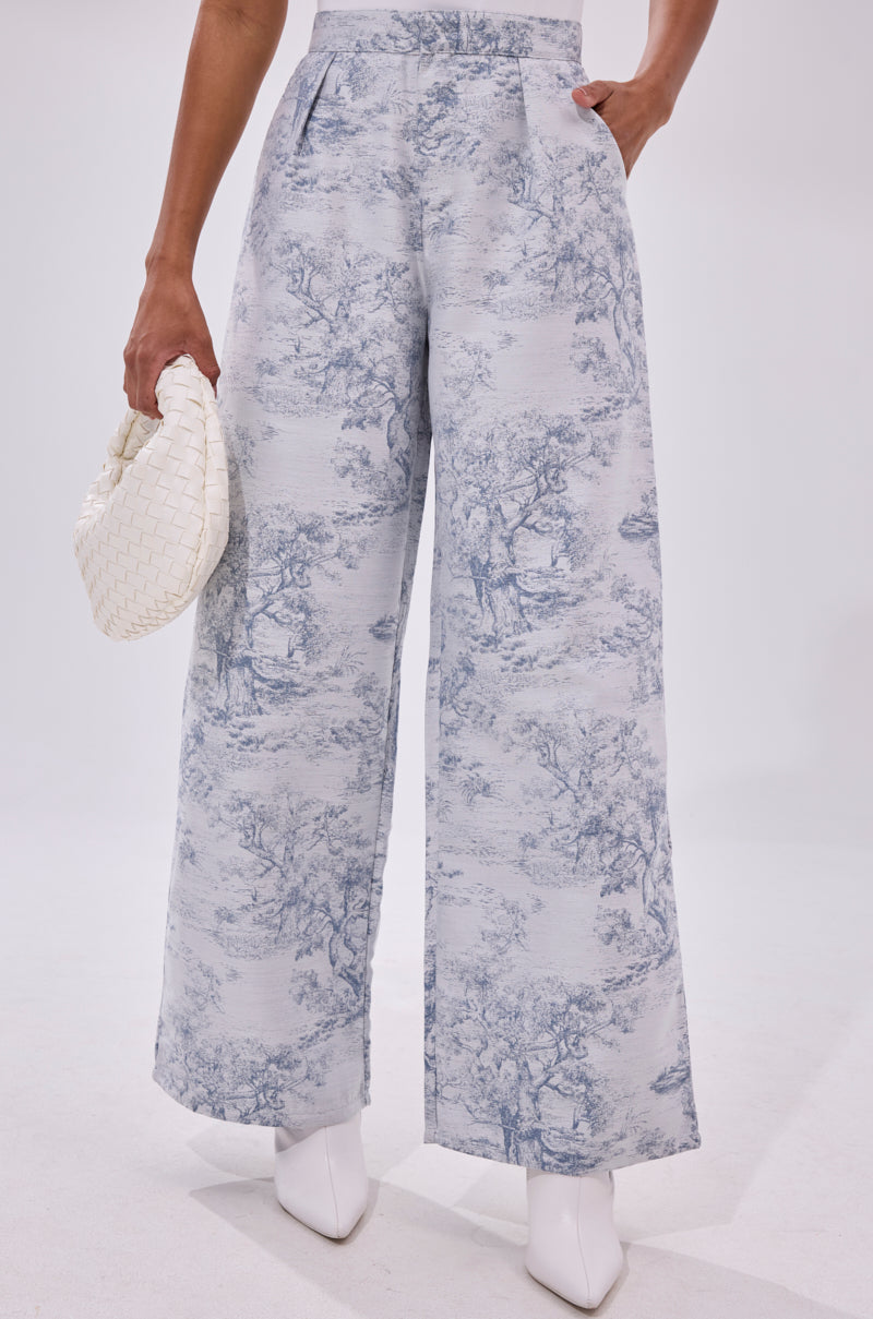 A person wears the BOOKED N BUSY JACQUARD TROUSER, featuring a scenic toile pattern, paired with white pointed-toe boots and a small woven white handbag against a plain white background.