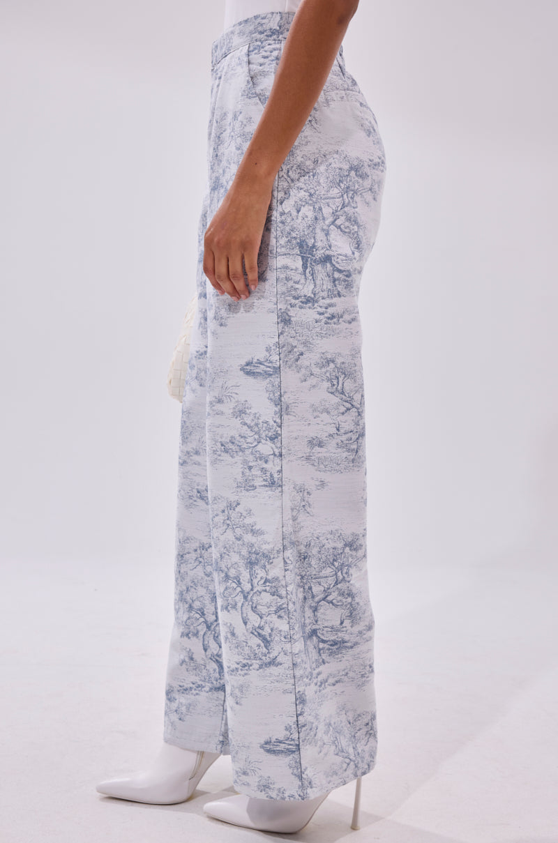 A person stands sideways against a plain white background, wearing BOOKED N BUSY JACQUARD TROUSER with a blue and white toile-inspired pattern, wide-leg silhouette, and high waist, paired with white heeled boots.