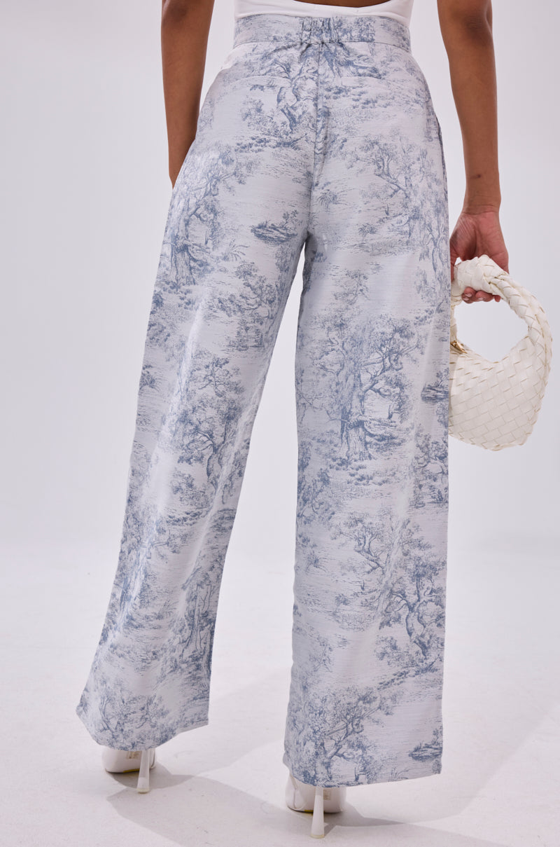 Woman models the BOOKED N BUSY JACQUARD TROUSER—high-rise wide-leg pants with a scenic toile pattern—holding a small white woven handbag, photographed from behind against a plain light background.