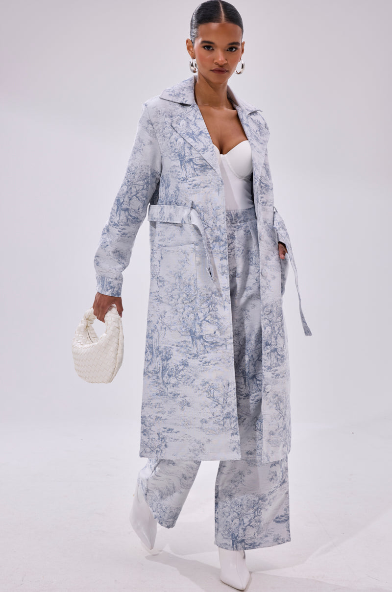 A woman poses confidently in BOOKED N BUSY JACQUARD TROUSER, a high-rise wide-leg pant with a light blue and white scenic toile pattern, paired with a matching trench coat, white top and boots, and carries a small white woven handbag.