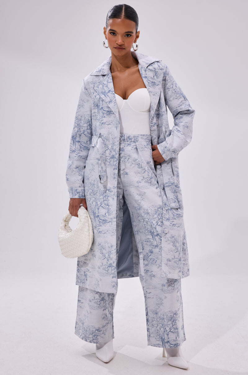 A woman poses confidently in the BOOKED N BUSY JACQUARD TROUSER—a light blue scenic toile high-rise wide-leg pant—paired with a matching trench coat, white bustier top, white heeled boots, hoop earrings, and a small white woven handbag.