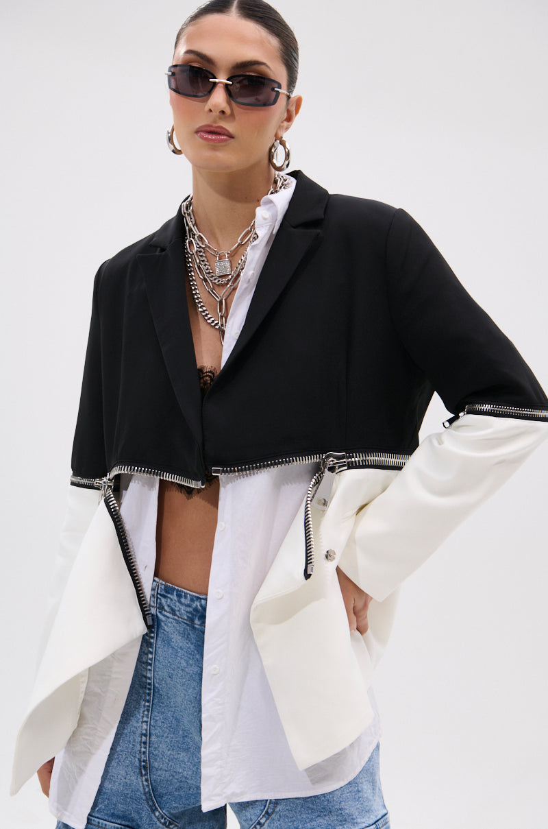 A woman in sunglasses wears layered silver chains, the DON'T TRIP ZIPPER BLAZER with a black and white colorblock design, an open-collar white shirt, and light blue jeans against a plain background.