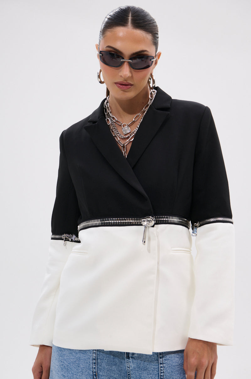 A woman in dark sunglasses, layered silver chain necklaces, and the DON'T TRIP ZIPPER BLAZER with a statement waist detail, paired with light blue jeans, stands against a plain white background.