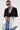 A woman stands confidently against a white background in the DON'T TRIP ZIPPER BLAZER, styled with dark sunglasses, layered chain necklaces, an unbuttoned white shirt, and blue jeans—a true statement piece.